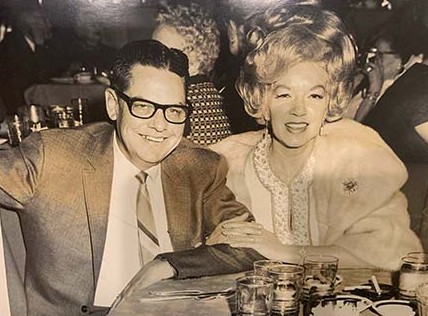 Black and white photo of Bill Barnes in glasses and a suit seated beside his wife