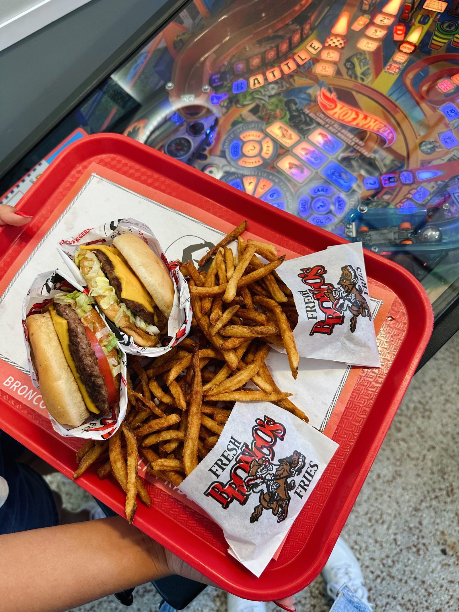 Two Bronco's burgers and a pile of hand-cut fries on a red tray, held over a pinball machine in the game room
